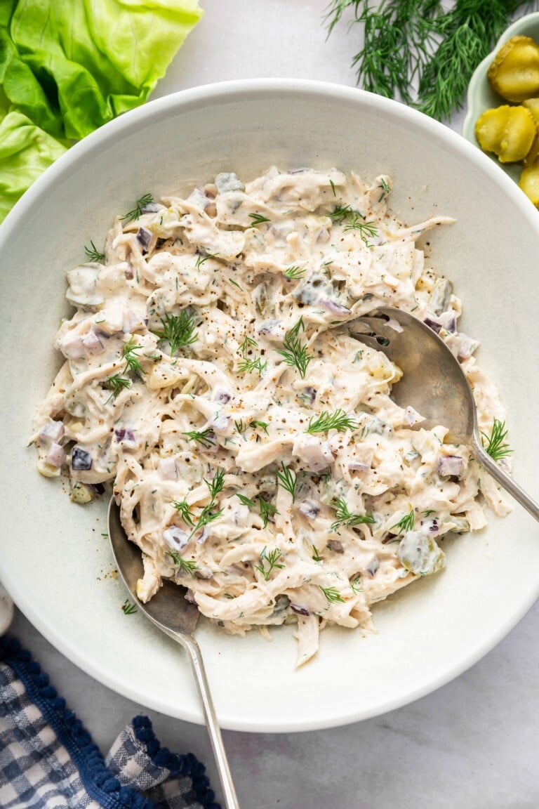 Dill pickle chicken salad in a large serving bowl with two serving spoons and topped with fresh dill.