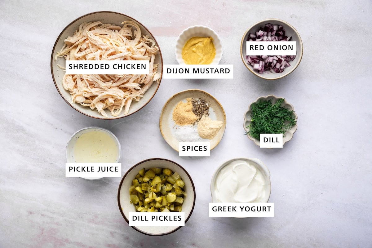 Ingredients measured out for dill pickle chicken salad: shredded chicken, dijon mustard, red onion, dill, greek yogurt, dill pickles, pickle juice, and spices.