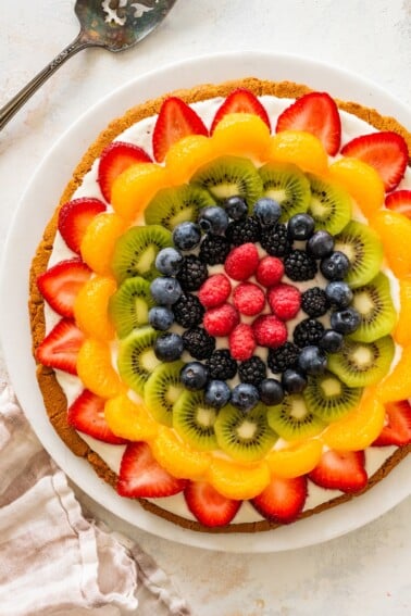 Sugar cookie fruit pizza topped with strawberries, mandarin oranges, kiwi, blueberries, blackberries and raspberries arranged in a colorful pattern.