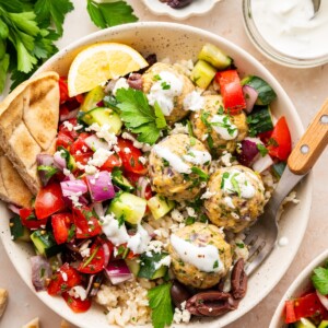 A serving of Greek turkey meatballs in a bowl with rice, a fresh vegetable salad, tzatziki sauce, pita bread, olives, lemon slices, and fresh herbs.