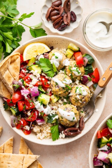 A serving of Greek turkey meatballs in a bowl with rice, a fresh vegetable salad, tzatziki sauce, pita bread, olives, lemon slices, and fresh herbs.