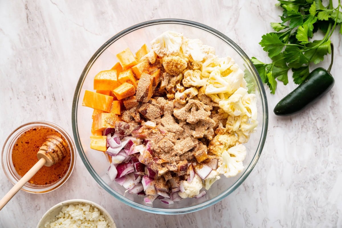 A large glass mixing bowl with red onion, cauliflower, sweet potato, and a spice blend.