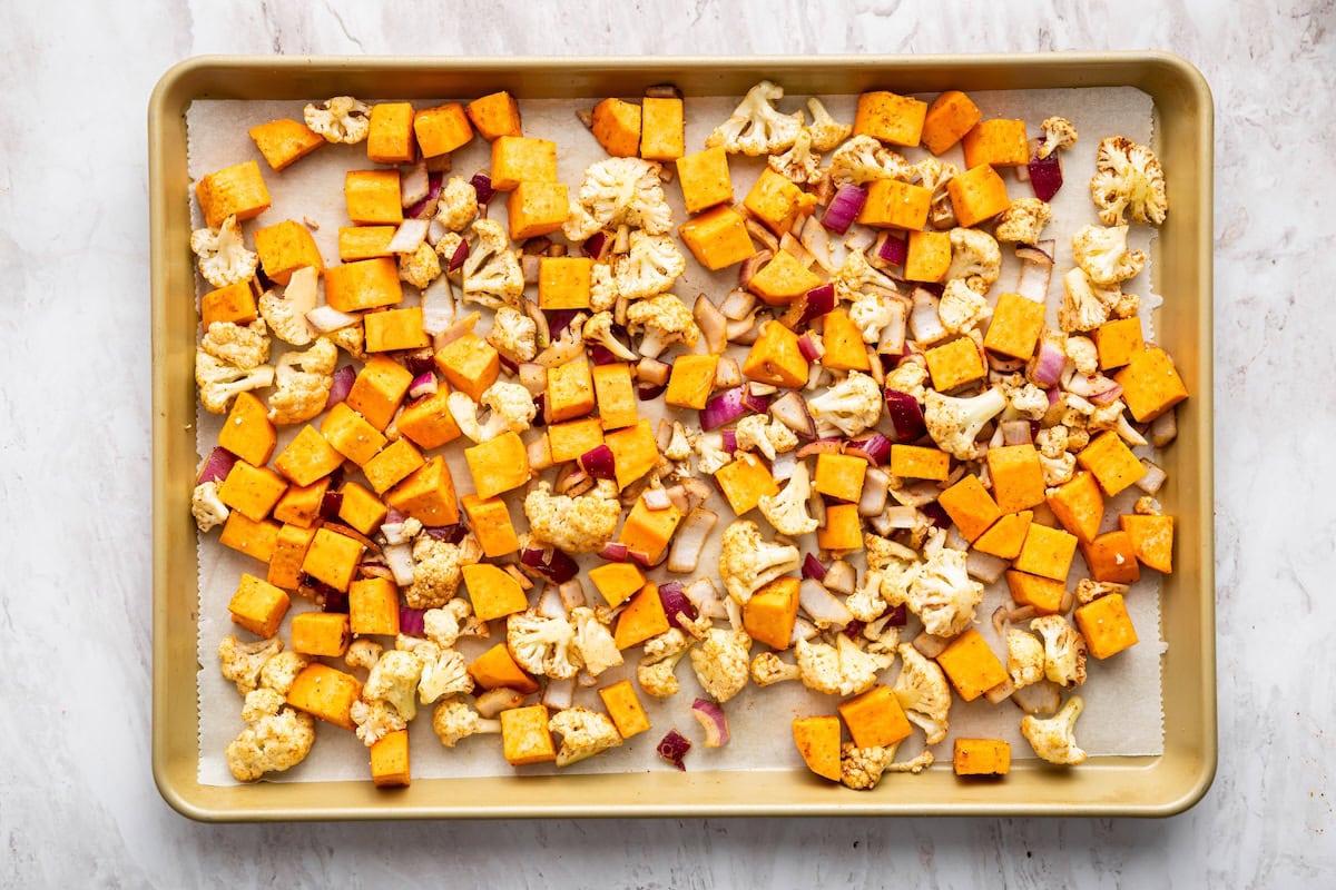 A large sheet pan with diced sweet potatoes, cauliflower florets, and diced red onion.