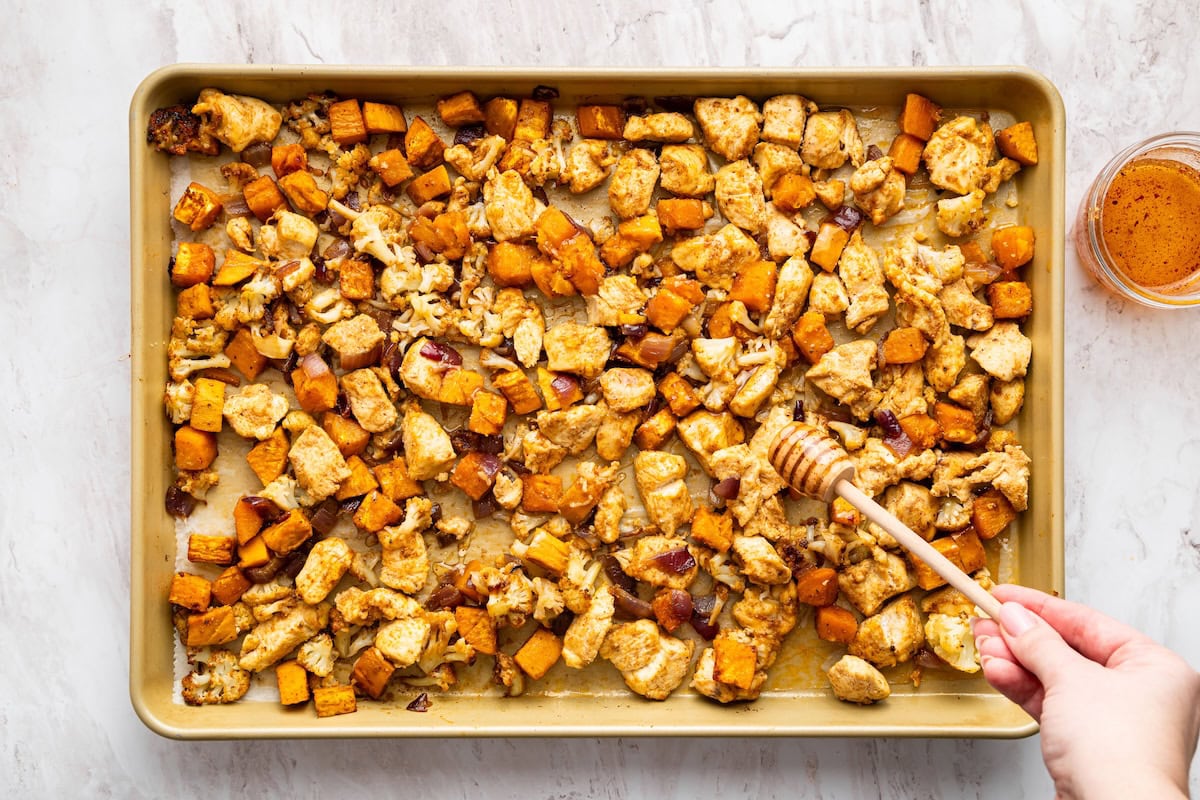A womans hand uses a honey dipper to drizzle hot honey over a sheet pan with roasted vegetables and chicken.