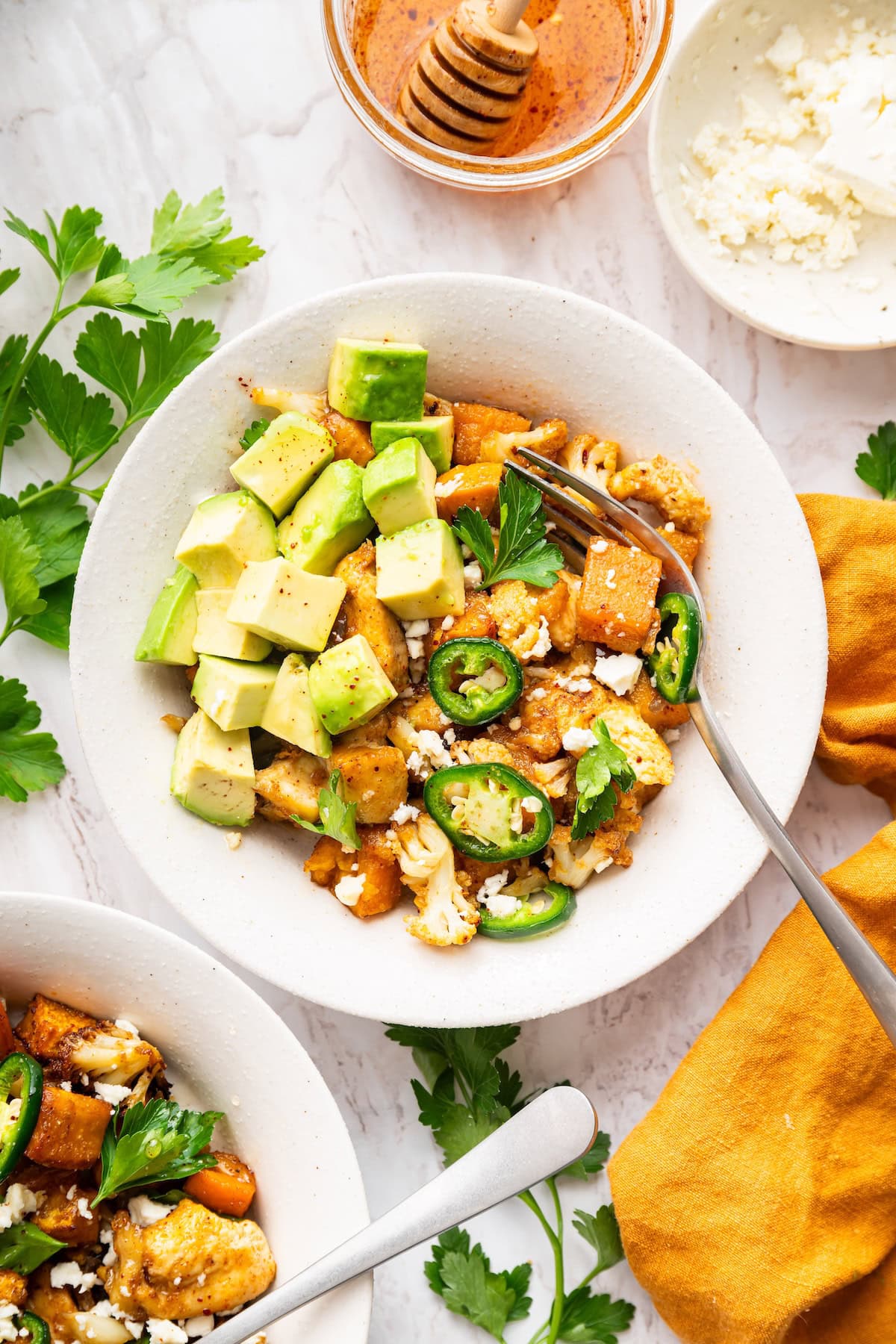 A serving of hot honey chicken sheet pan meal in a bowl topped with avocado chunks, sliced jalape&ntilde;o, fresh herbs, and feta cheese.