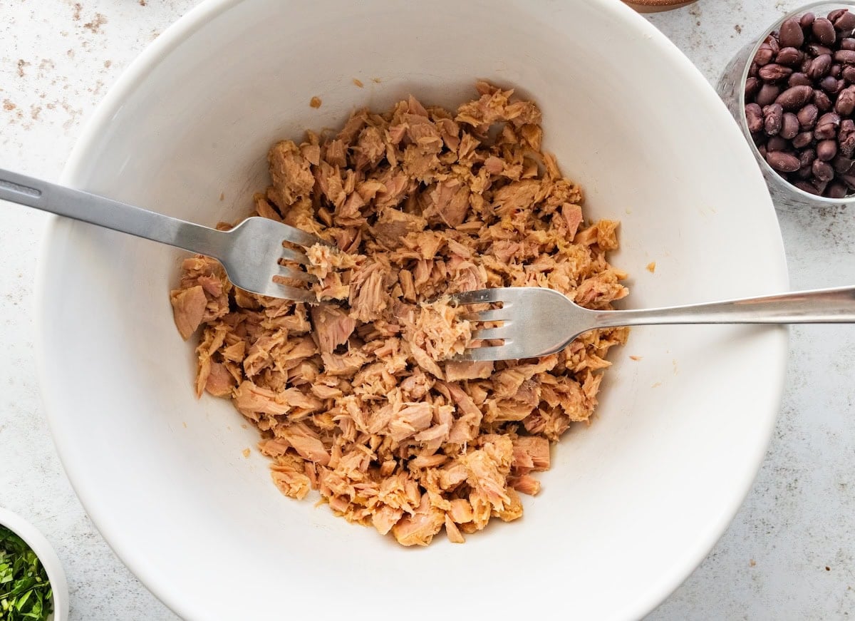 Canned tuna is shredded with two forks in a large bowl.