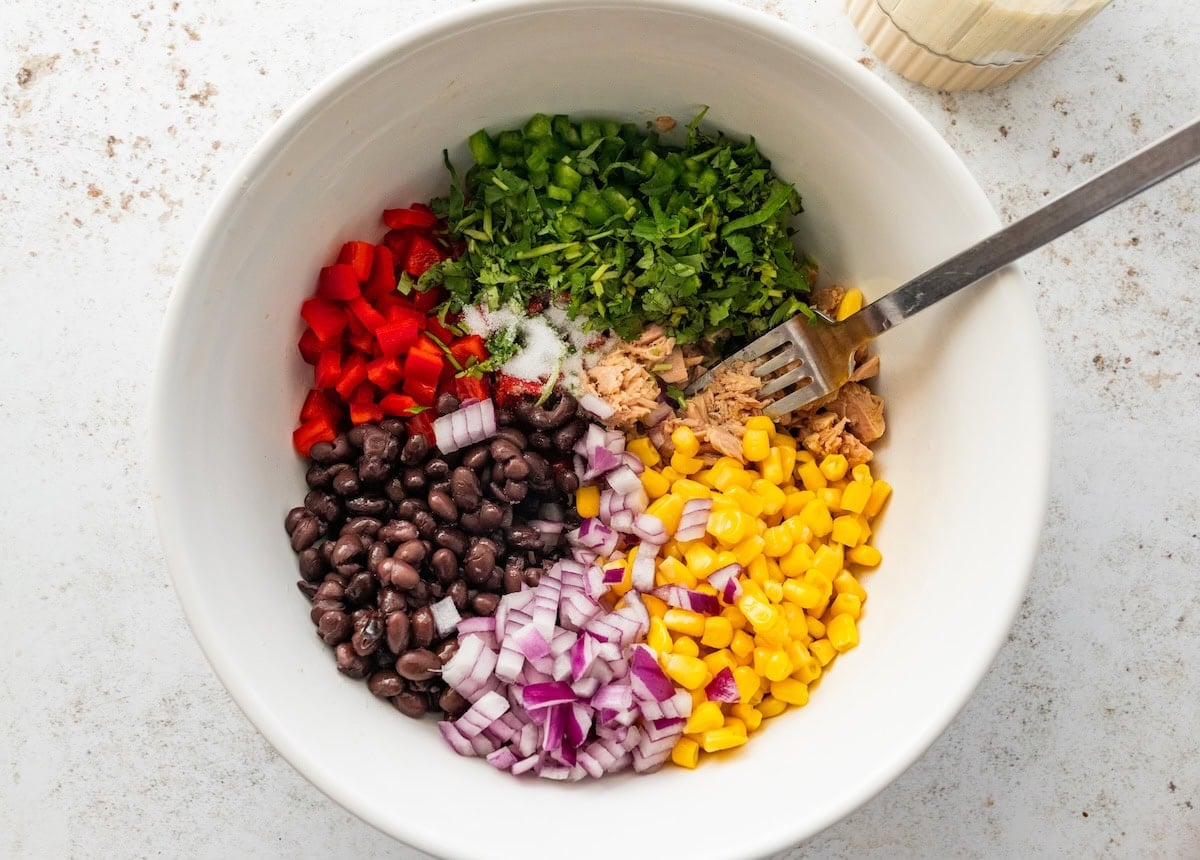 A large mixing bowl with canned tuna, jalapeño, red bell pepper, black beans, red onion, and corn.