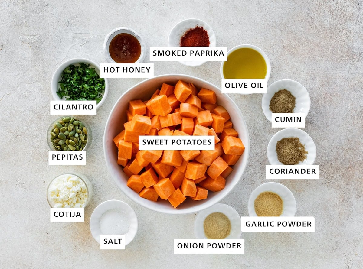 Ingredients measured out for roasted Mexican sweet potatoes: Sweet potatoes, smoked paprika, olive oil, cumin, coriander, garlic powder, onion powder, salt, cotija, pepitas, cilantro, and hot honey.