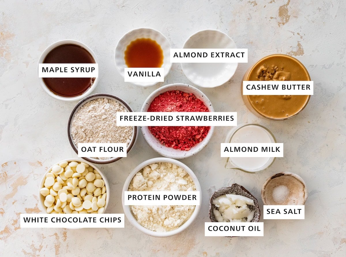 Ingredients for strawberry protein balls: maple syrup, oat flour, white chocolate chips, protein powder, coconut oil, sea salt, almond milk, cashew butter, almond extract and vanilla extract.