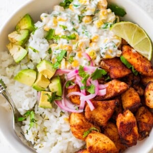 Seasoned chicken, Mexican street corn, cilantro lime rice, avocado, pickled red onion, and a lime wedge in a bowl.