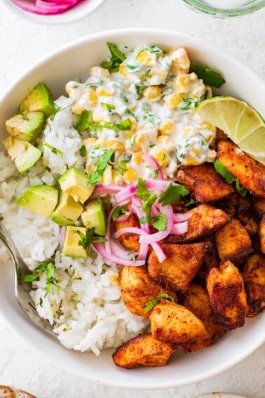 Seasoned chicken, Mexican street corn, cilantro lime rice, avocado, pickled red onion, and a lime wedge in a bowl.