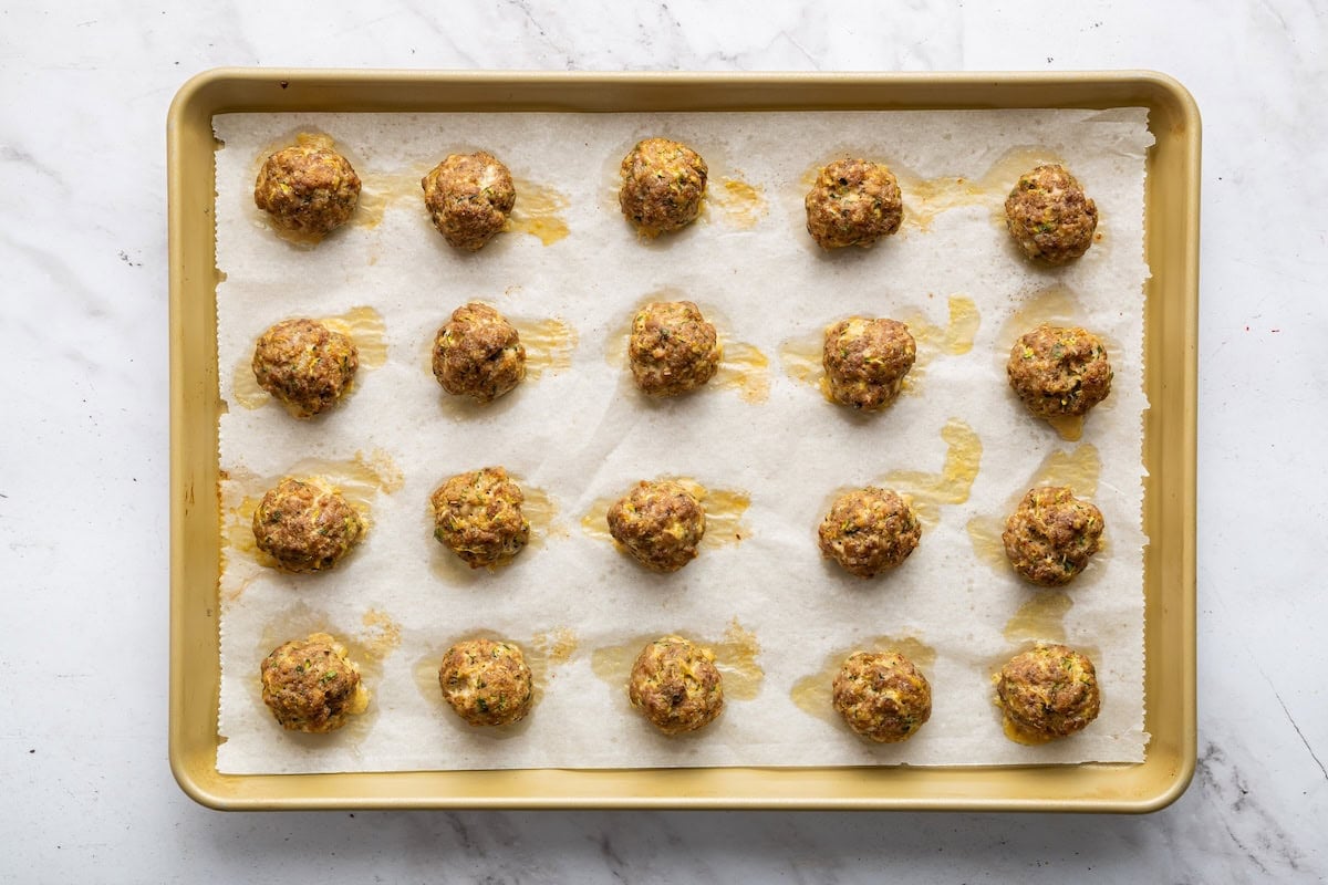 Zucchini meatballs spread out on a baking tray after being baked in the oven.