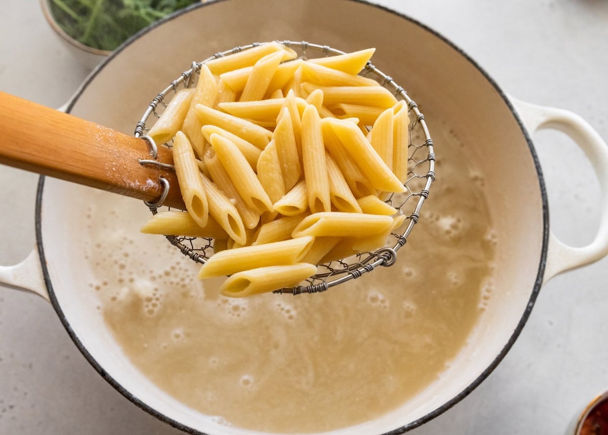Cooked pasta held over a pot of boiling water by a strainer spoon.
