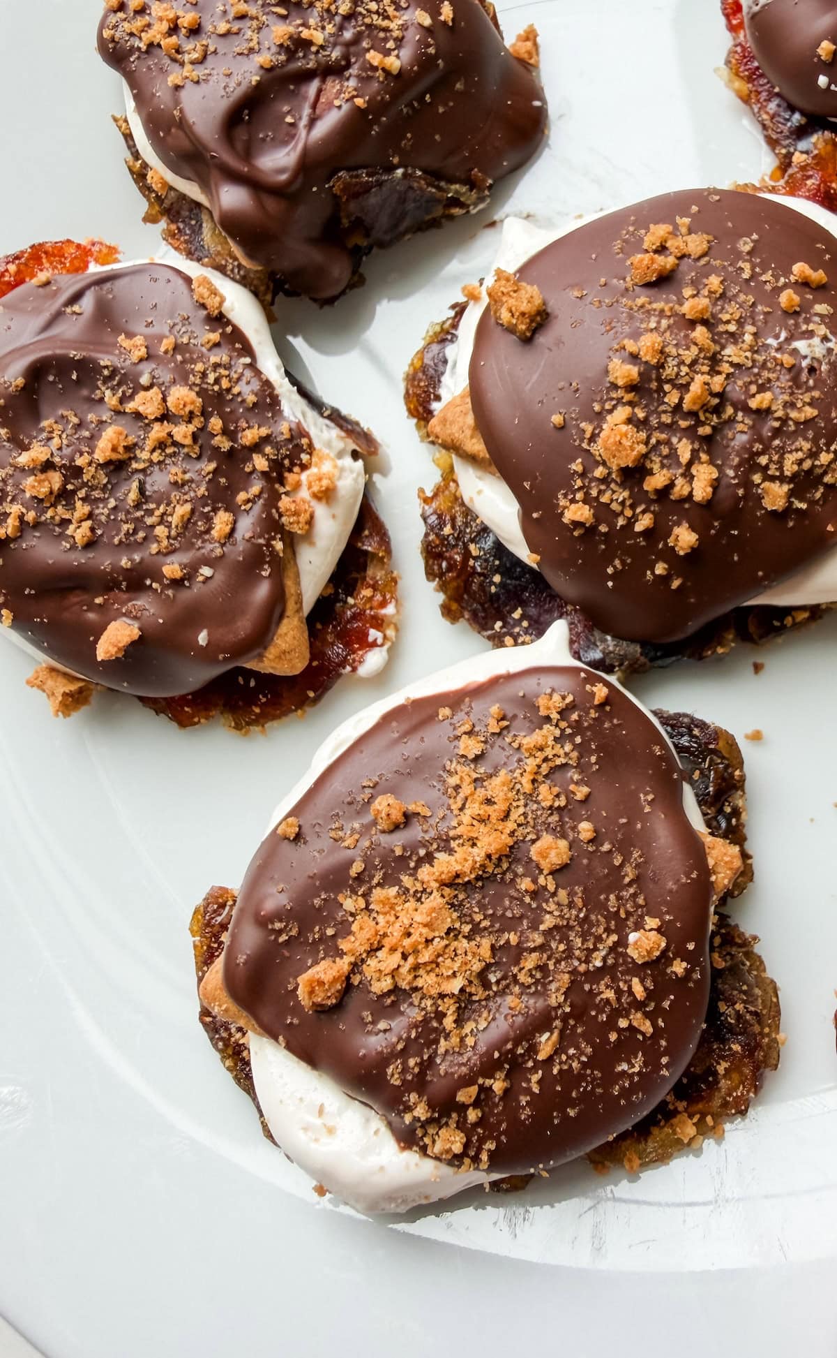 Smashed date s'mores on a white plate topped with melted chocolate and crushed graham crackers.