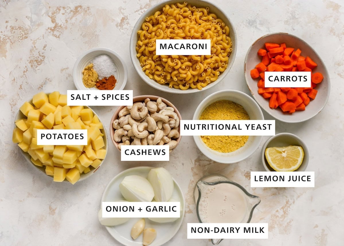 Ingredients measured out in bowls and on plates to make vegan mac and cheese: carrots, lemon, nutritional yeast, elbow macaroni, non-dairy milk, cashews, garlic, onion, salt, turmeric, mustard powder and potatoes.