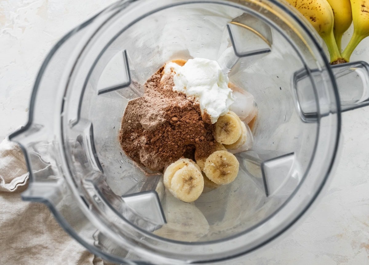 Protein powder, frozen banana, yogurt, cacao powder, milk, and vanilla in a blender.