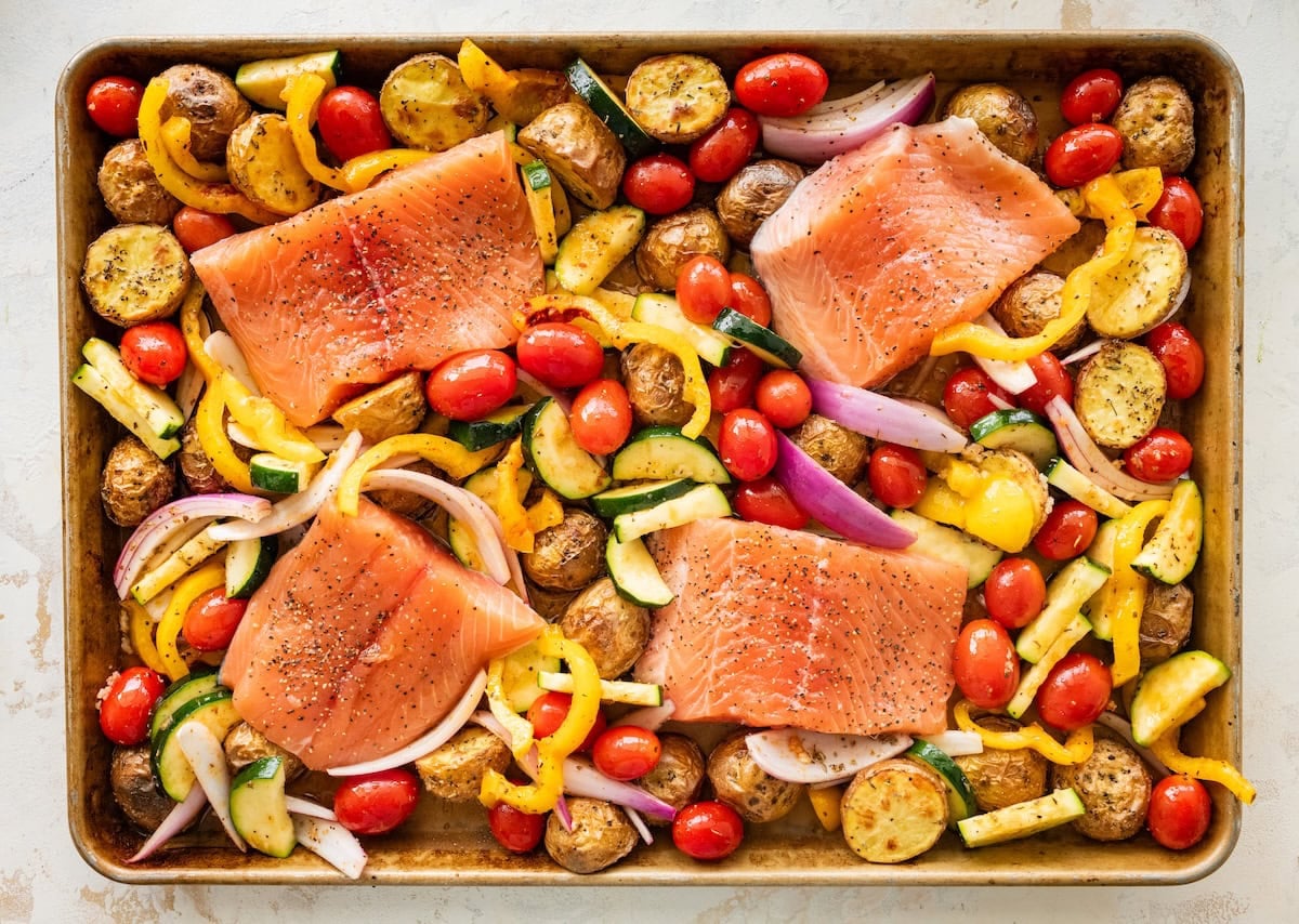 Roasted potatoes and veggies on a baking tray with four raw salmon fillets.
