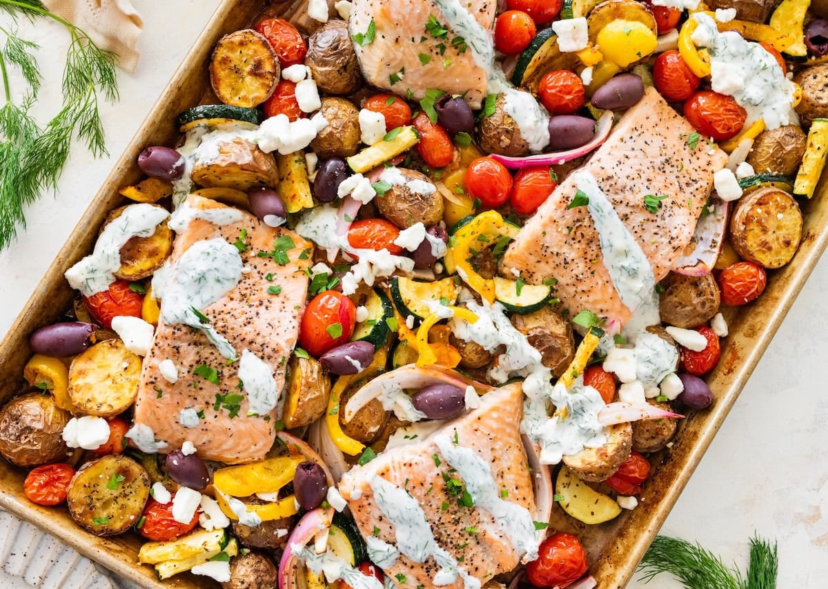 Salmon sheet pan meal with roasted veggies and a dill yogurt sauce on a baking tray.