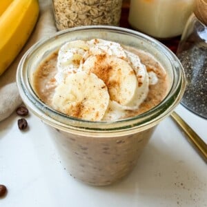 Banana bread latte overnight oats in a glass jar topped with whipped cream, banana slices and cinnamon.