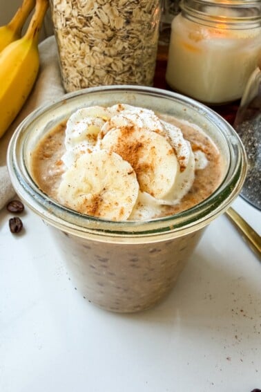 Banana bread latte overnight oats in a glass jar topped with whipped cream, banana slices and cinnamon.