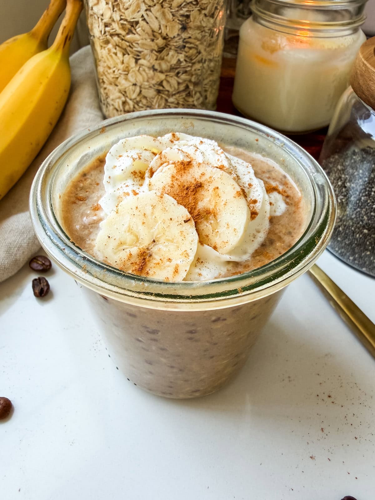 Banana bread latte overnight oats in a glass jar topped with whipped cream, banana slices and cinnamon.