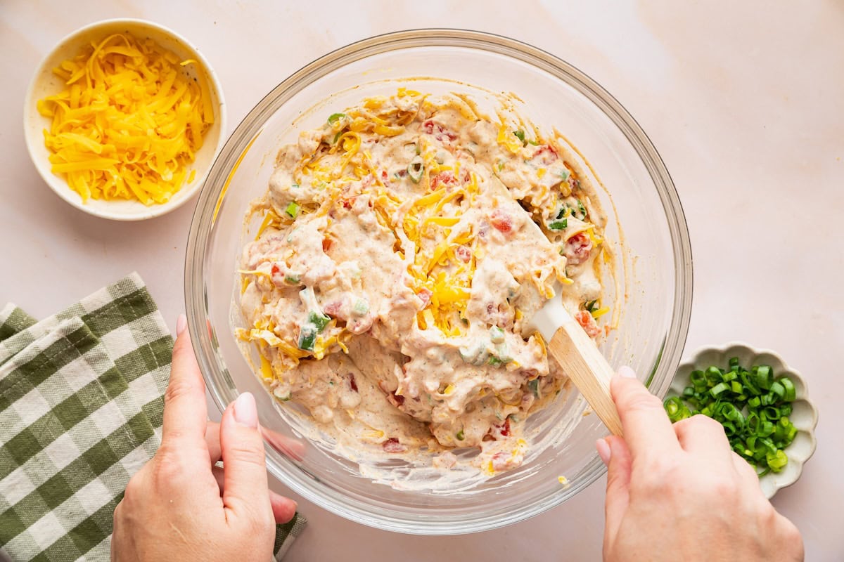 A womans hand uses a spatula to combine ingredients in a large glass bowl for a cottage cheese dip.