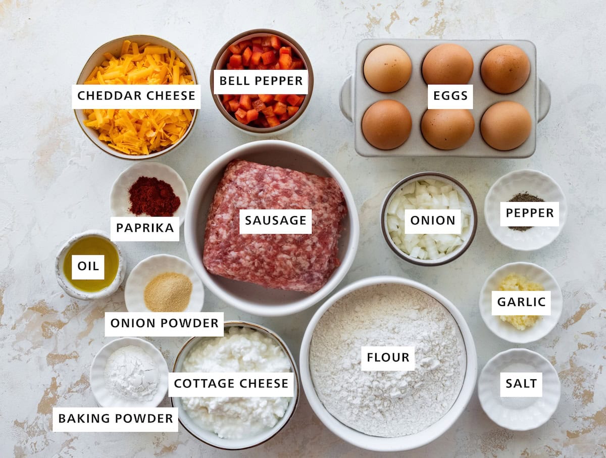 Breakfast biscuit ingredients arranged in labeled bowls, including flour, eggs, sausage, cottage cheese, cheddar, vegetables and seasonings.