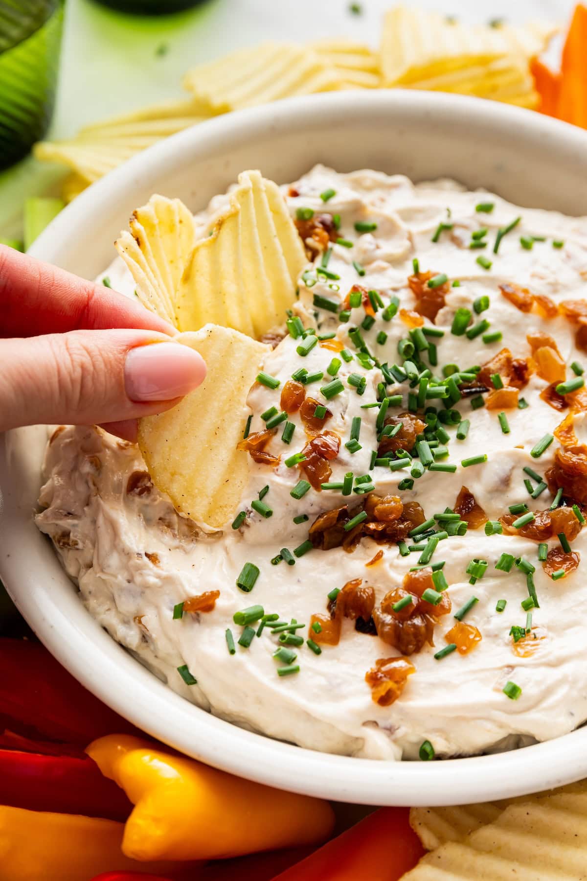 A woman's hand dipping a chip in French onion dip. The dip is topped with caramelized onions and fresh chives.