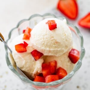 Scoops of greek yogurt ice cream in a glass bowl topped with chopped strawberries and served with a spoon.