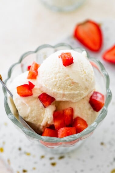 Scoops of greek yogurt ice cream in a glass bowl topped with chopped strawberries and served with a spoon.