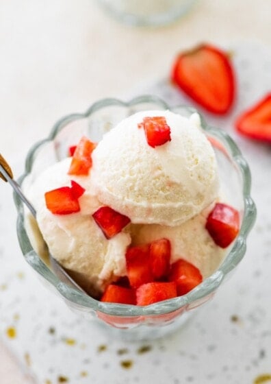 Scoops of greek yogurt ice cream in a glass bowl topped with chopped strawberries and served with a spoon.