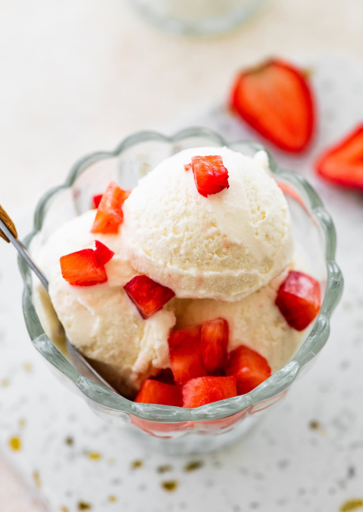 Scoops of greek yogurt ice cream in a glass bowl topped with chopped strawberries and served with a spoon.