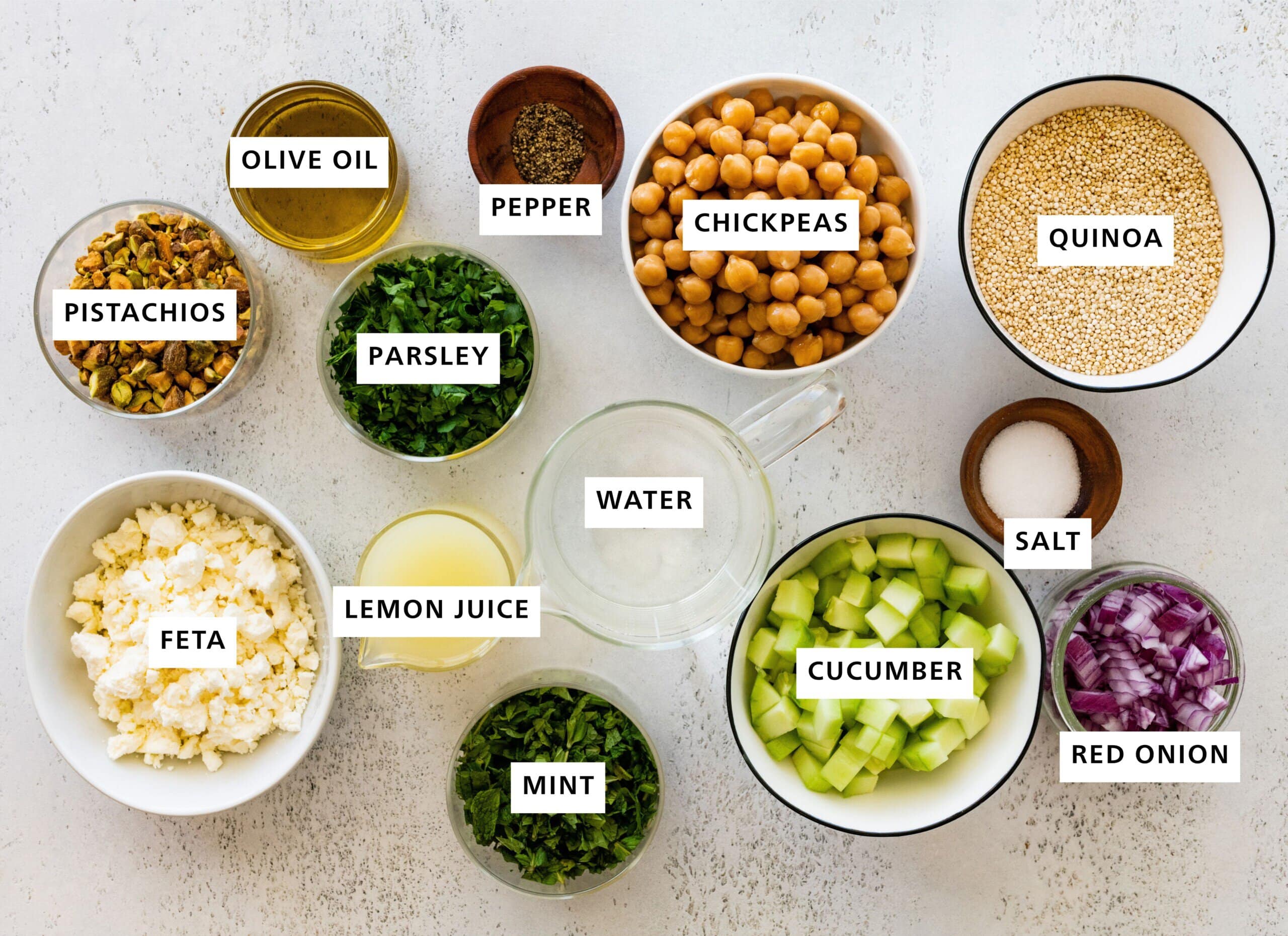 Ingredients measured out to make the Jennifer Aniston salad: salt, pepper, mint, quinoa, chickpeas, red onion, lemon juice, pistachios, feta, parsley, cucumber, water and olive oil.