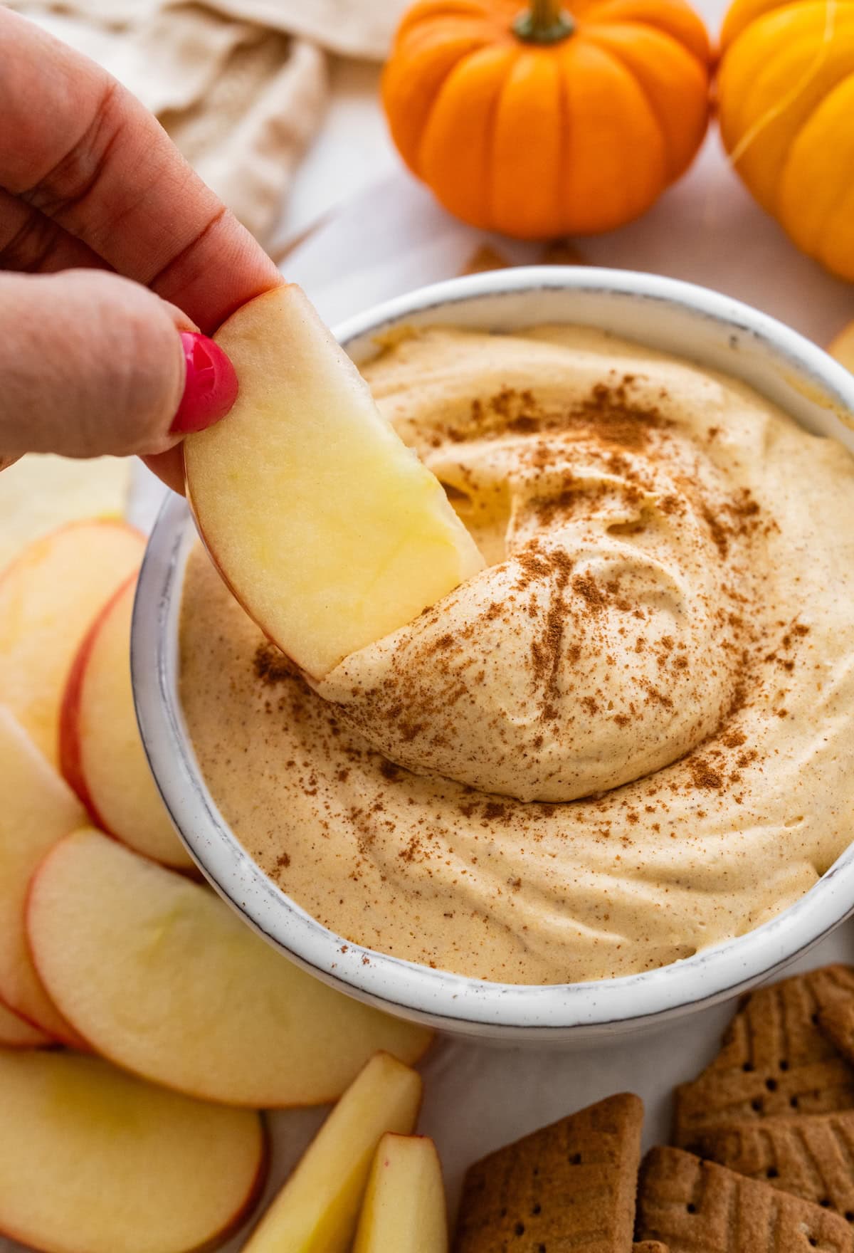A womans hand dipping an apple slice into a small bowl of pumpkin fluff dip.