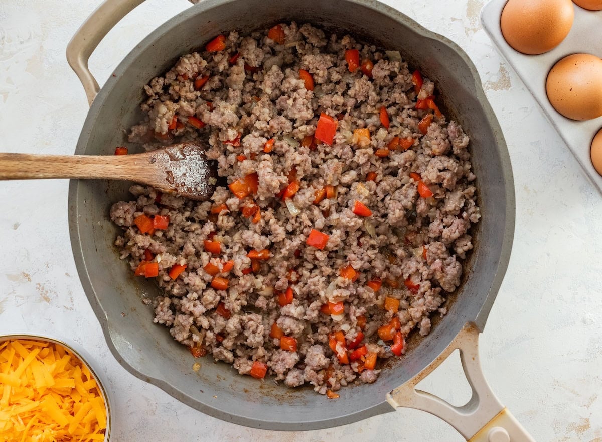 Crumbled sausage cooking with diced red bell pepper and onion in a skillet.