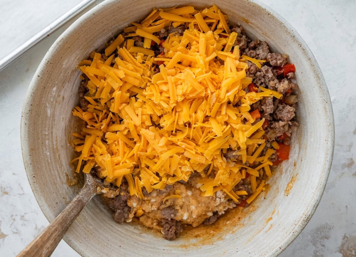 Shredded cheddar and cooked sausage mixture added to breakfast biscuit batter in a mixing bowl.