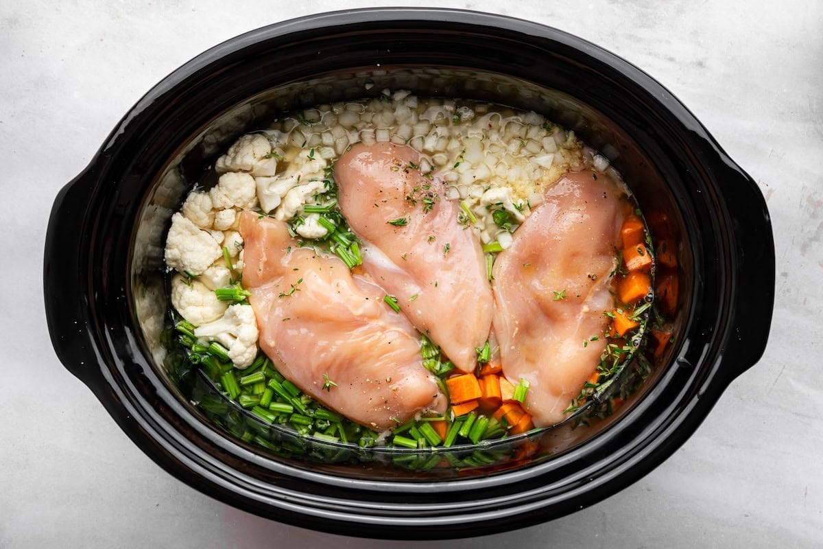 Raw chicken breasts, cauliflower, carrots, celery and herbs in a slow cooker before cooking.