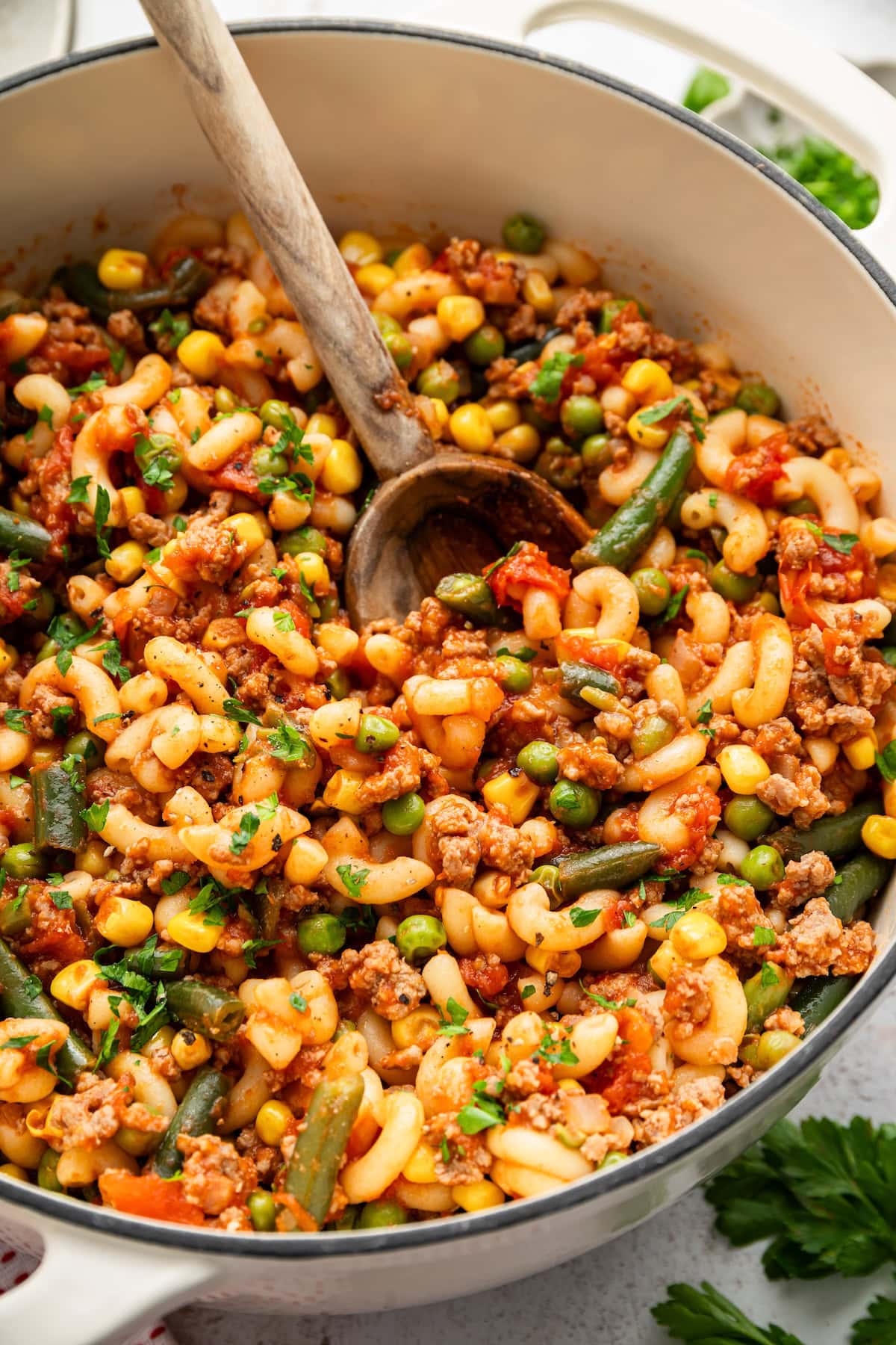 Close-up of beef goulash with macaroni, corn, peas and green beans beside a wooden spoon.