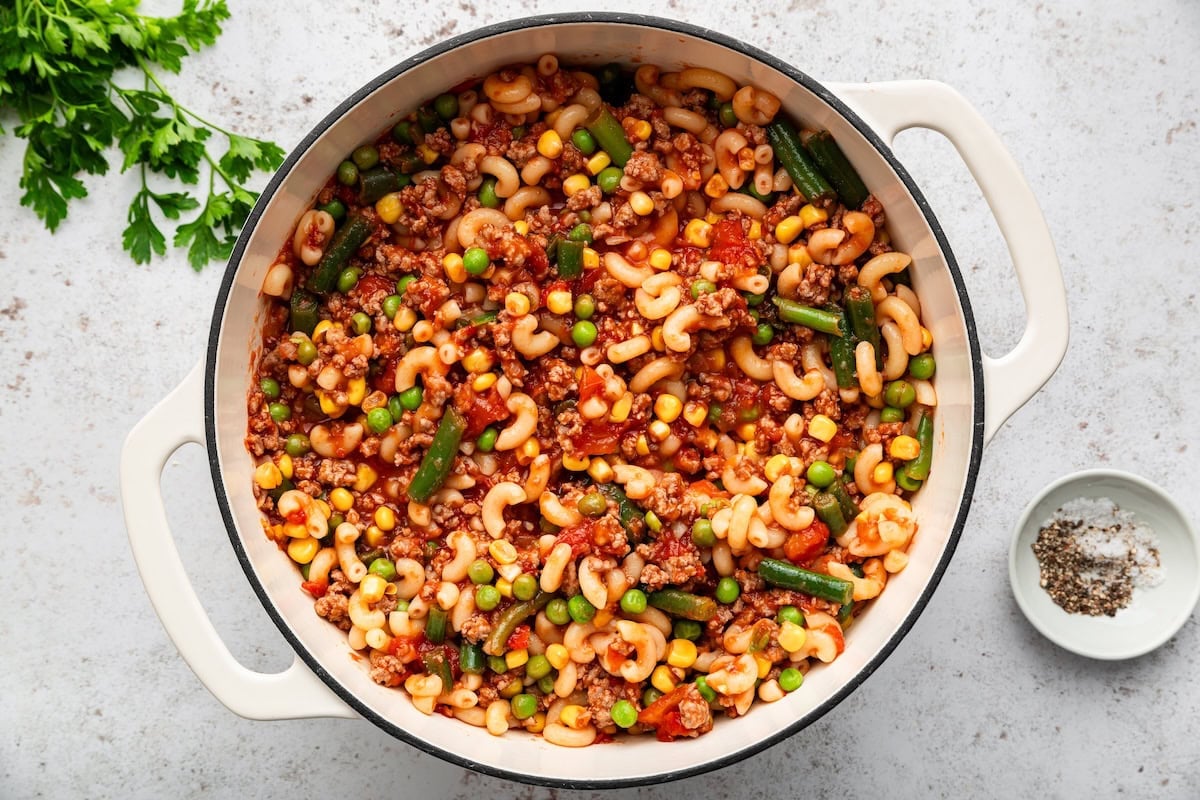 Cooked elbow macaroni mixed with beef, tomatoes, corn, peas and green beans in a Dutch oven.