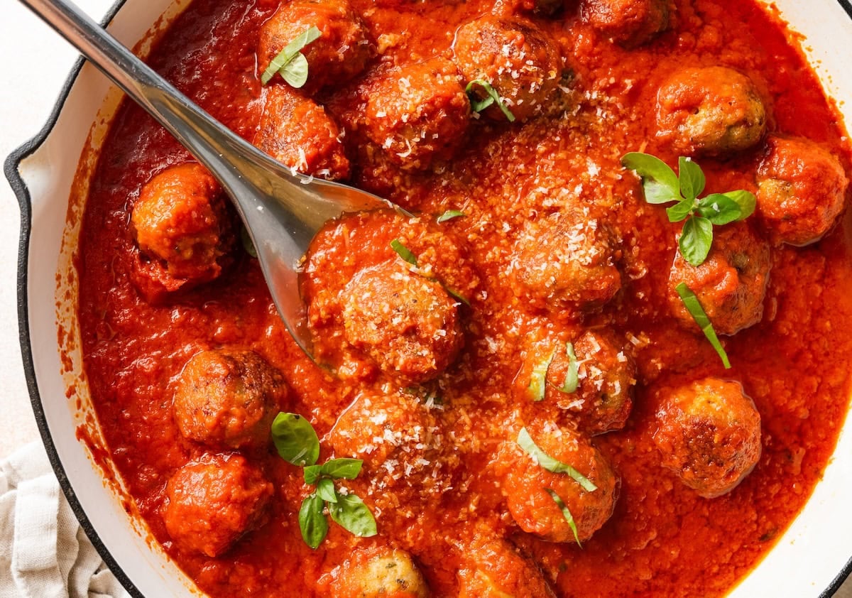 Spoon lifting a chicken meatball from a skillet of marinara sauce.