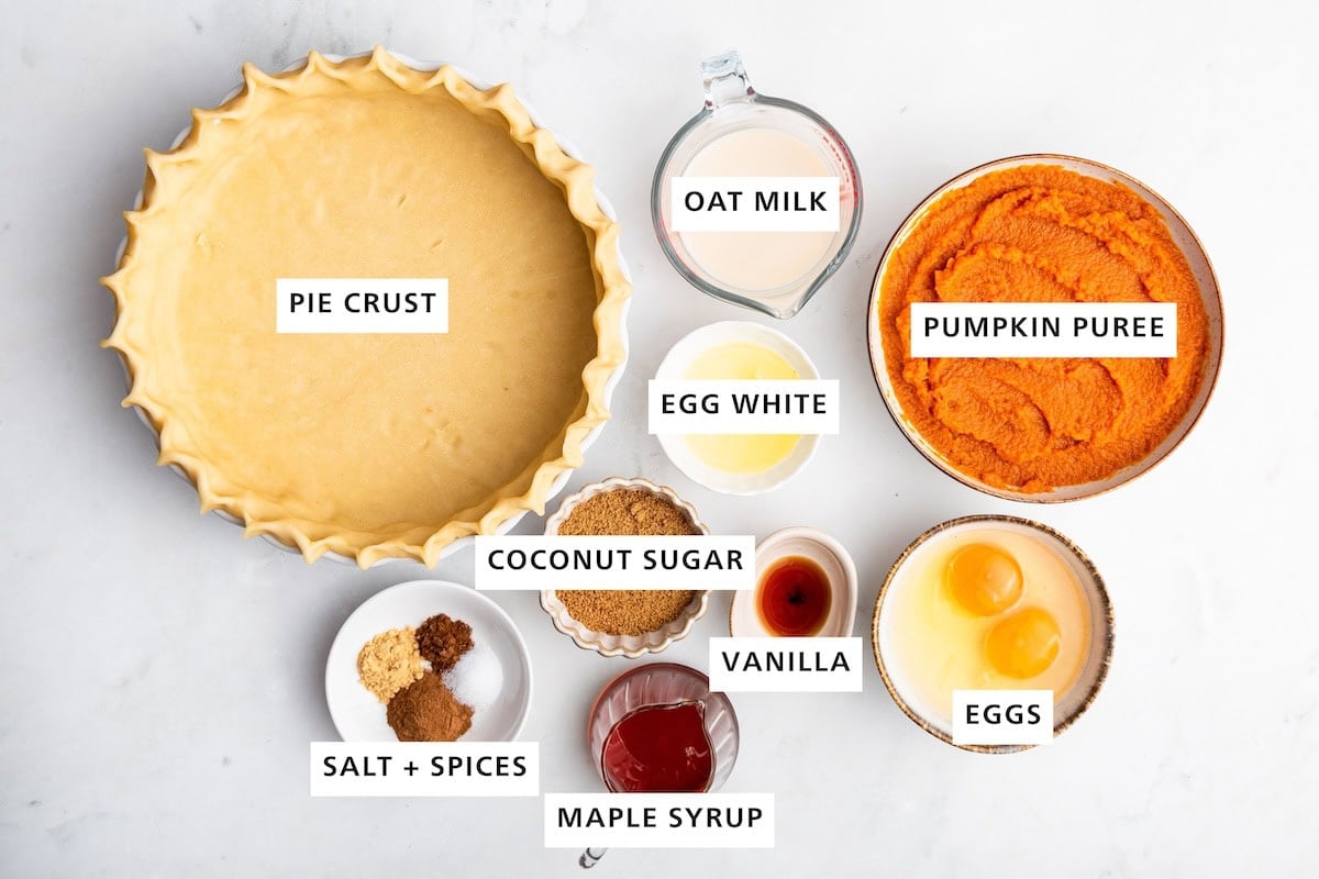Ingredients measured out for healthy pumpkin pie include: pie crust, oat milk, pumpkin puree, egg white, coconut sugar, salt, spices, maple syrup, vanilla, and eggs.