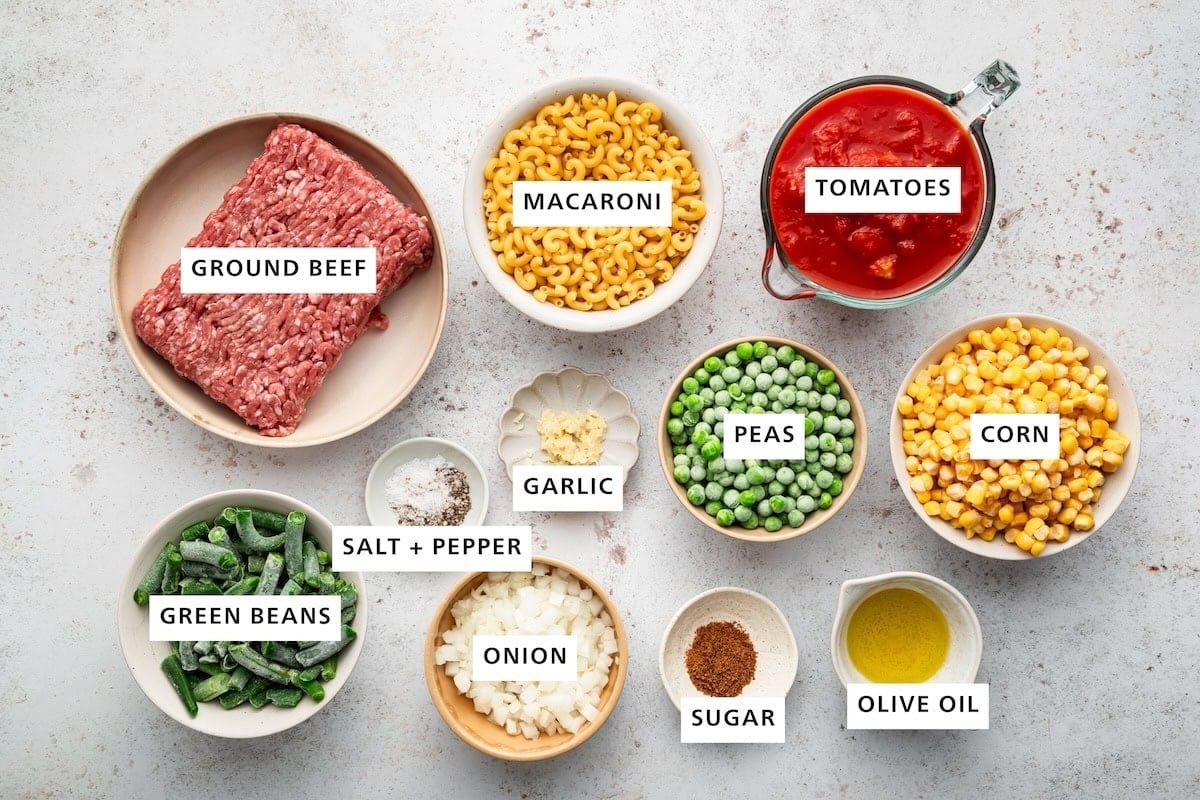 Ingredients measured out for mom's goulash recipe includes: ground beef, macaroni, tomatoes, corn, peas, garlic, salt, pepper, green beans, onion, sugar, and olive oil.