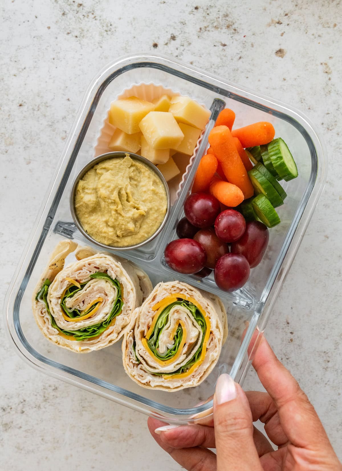 Glass container with turkey pinwheel sandwiches, hummus, cubed cheese, red grapes, baby carrots and sliced cucumbers.