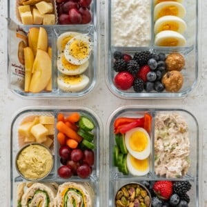 Four high protein snack boxes with eggs, cottage cheese, turkey pinwheels, chicken salad, fruit and veggies in glass meal prep containers.