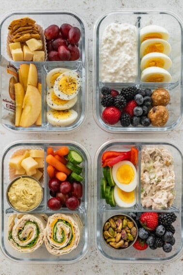 Four high protein snack boxes with eggs, cottage cheese, turkey pinwheels, chicken salad, fruit and veggies in glass meal prep containers.