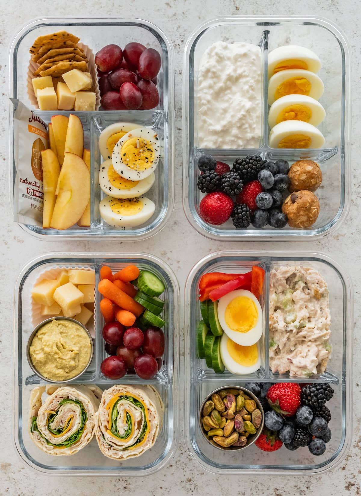 Four high protein snack boxes with eggs, cottage cheese, turkey pinwheels, chicken salad, fruit and veggies in glass meal prep containers.