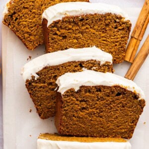Overhead view of a partially sliced pumpkin bread loaf topped with cottage cheese frosting, beside cinnamon sticks.