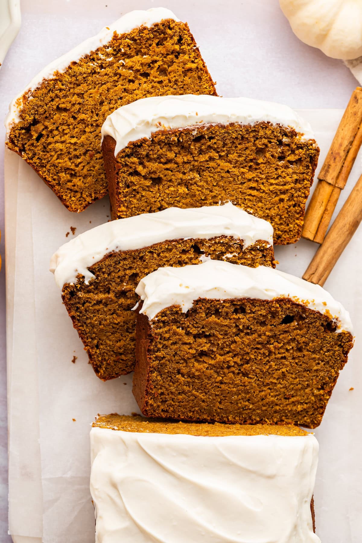 Overhead view of a partially sliced pumpkin bread loaf topped with cottage cheese frosting, beside cinnamon sticks.