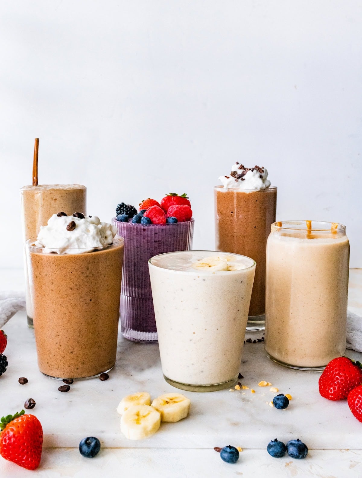 Overhead view of six protein shakes with fresh berries, sliced banana, peanut butter drizzle and whipped cream with chocolate pieces or coffee beans.