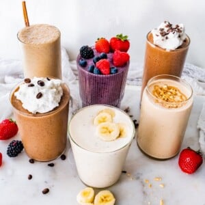Six protein shakes arranged on a marble board, topped with berries, banana slices, peanut butter, chopped peanuts and whipped cream.